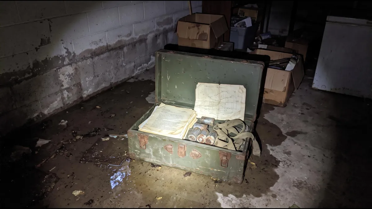 An old green military footlocker, open on a basement floor, lit by a flashlight